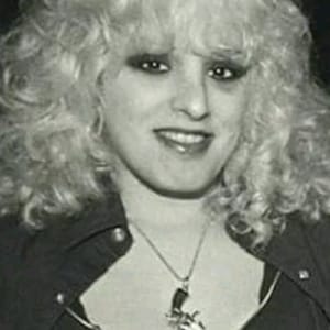 May include: A black and white photo of a woman with blonde curly hair. She is wearing a black leather jacket with a zipper and a silver necklace with a gun pendant.