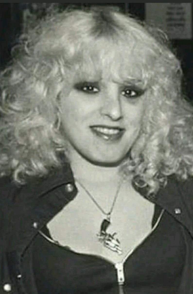 May include: A black and white photo of a woman with blonde curly hair. She is wearing a black leather jacket with a zipper and a silver necklace with a gun pendant.