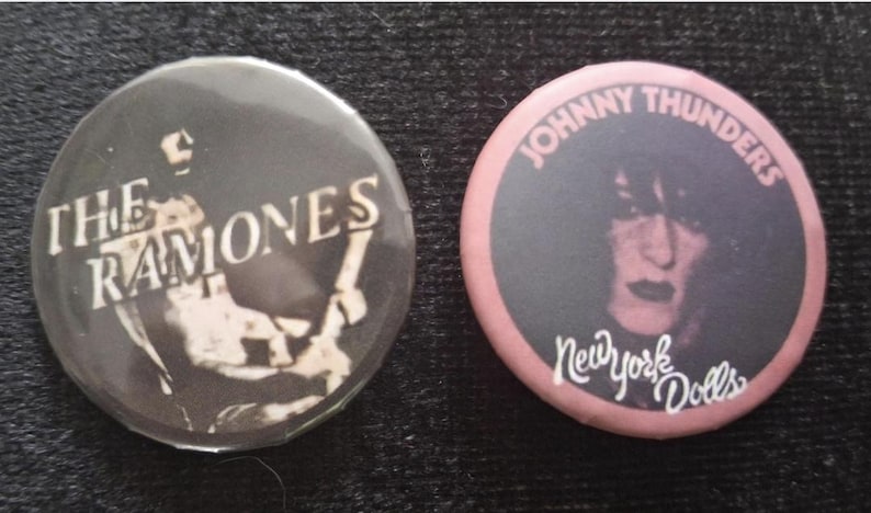 May include: Two round pinback buttons. One is black with the text "THE RAMONES" and a band image. The other is pink with "JOHNNY THUNDERS" and "New York Dolls" text, plus a portrait.