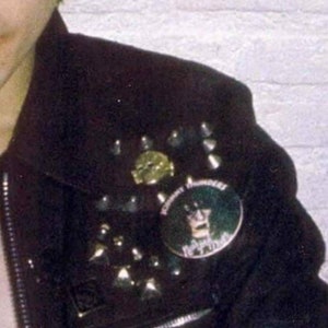 May include: Black leather jacket adorned with silver studs and a round button featuring a graphic. The jacket has a zipper and a collar. The button has the text "Johnny Thunders" and a graphic of a person.