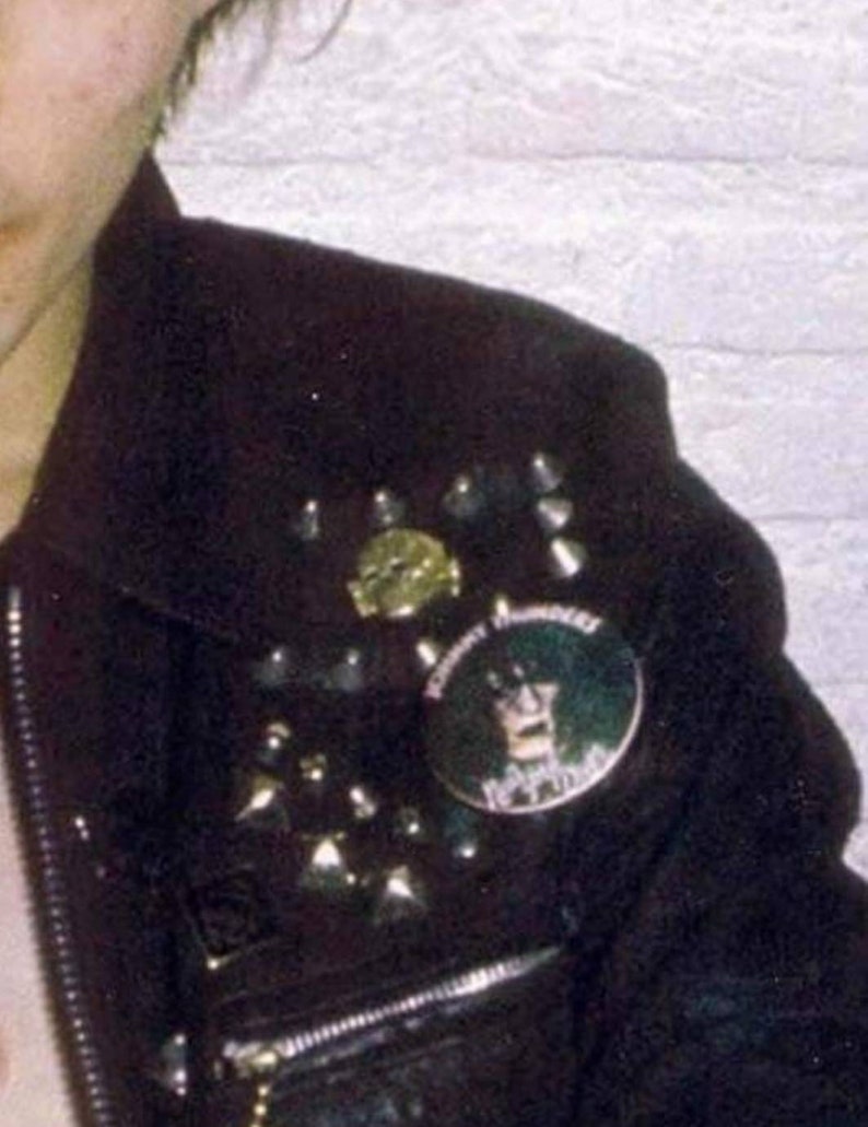 May include: Black leather jacket adorned with silver studs and a round button featuring a graphic. The jacket has a zipper and a collar. The button has the text "Johnny Thunders" and a graphic of a person.