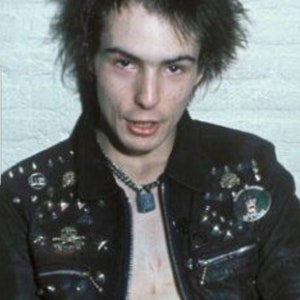 May include: A portrait of a person with spiky black hair and a black leather jacket adorned with metal studs and pins. A silver chain necklace with a pendant is visible. The person is looking directly at the camera.