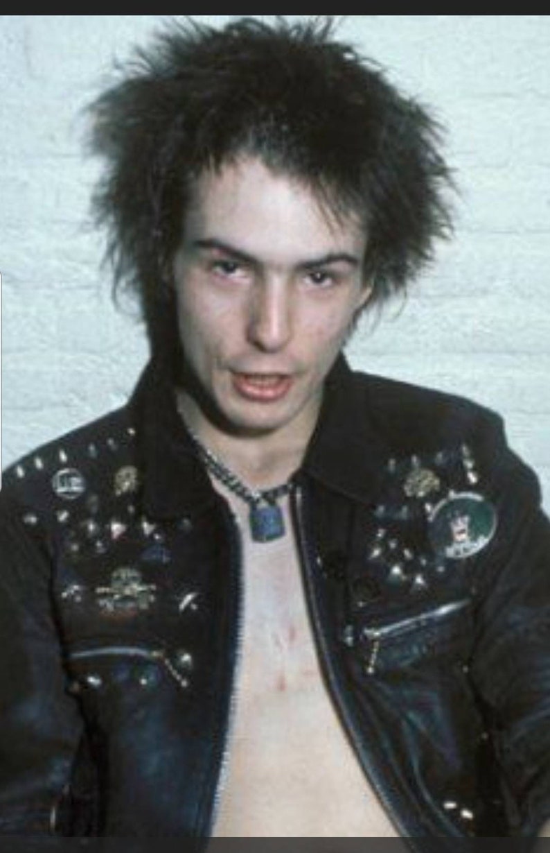 May include: A portrait of a person with spiky black hair and a black leather jacket adorned with metal studs and pins. A silver chain necklace with a pendant is visible. The person is looking directly at the camera.
