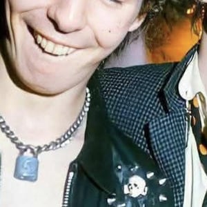 May include: A person smiling with closed eyes, wearing a silver chain necklace with a blue padlock pendant. The person is wearing a black leather jacket with patches and studs over a black and white checkered shirt.