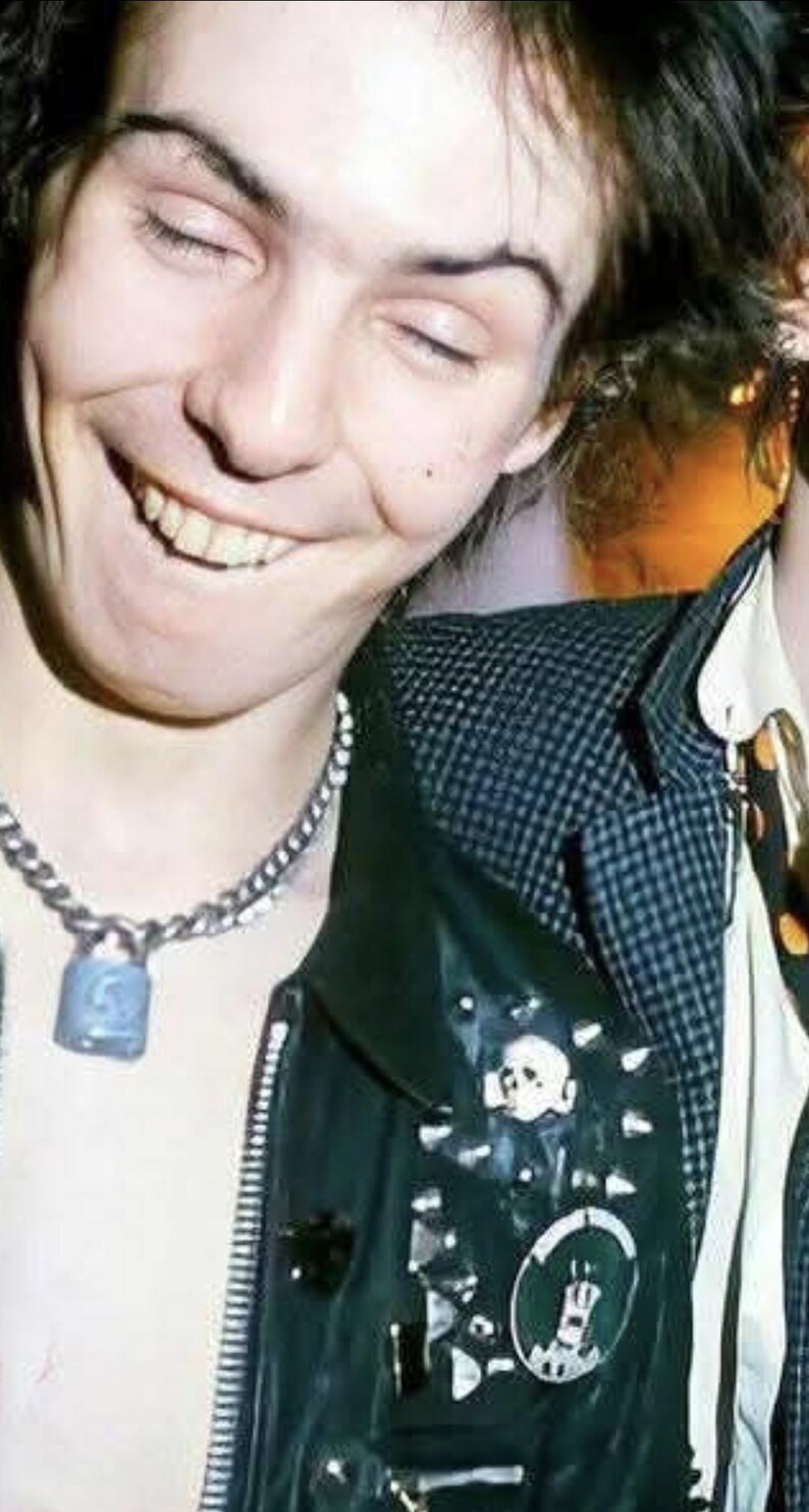 May include: A person smiling with closed eyes, wearing a silver chain necklace with a blue padlock pendant. The person is wearing a black leather jacket with patches and studs over a black and white checkered shirt.
