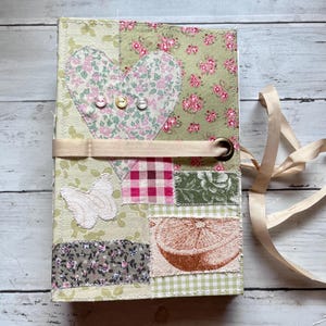 May include: A handmade journal with a patchwork design. The cover features a heart, butterfly, and orange slice appliqués, with floral and gingham patterns in shades of green, pink, and cream. A beige ribbon wraps around the journal.