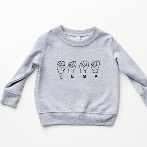 May include: Grey long-sleeved sweatshirt with the name "EMMA" spelled out in British Sign Language hand gestures.