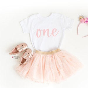 Peut inclure: Une jupe tutu rose, un t-shirt blanc avec le mot "one" en écriture rose, une paire de chaussures licorne roses et un bandeau licorne rose et or.