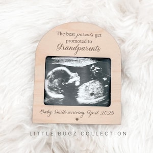 May include: Wooden frame with a white background and a black and white ultrasound image inside. The frame has the text "The best parents get promoted to grandparents" and "Baby Smith arriving April 2025" with a small heart symbol. The bottom of the frame has the text "LITTLE BUGZ COLLECTION".