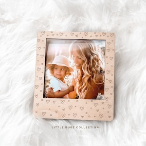 May include: A light wood picture frame with a heart pattern around the border. The frame has a clear plastic insert and a photo of a woman and child inside. The frame has the text "LITTLE BUGZ COLLECTION" printed on the bottom.