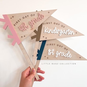 Könnte beinhalten: Holz-Wimpel-Flaggen für den ersten Schultag mit den Aufschriften "4th grade", "Kindergarten" und "1st grade". Jede Flagge hat eine andere farbliche Akzentuierung und wird von einem Holzstab gehalten. Der Text "LITTLE BUGZ COLLECTION" ist ebenfalls sichtbar.