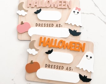 Puede incluir: Dos letreros de madera con temática de Halloween con la palabra "HALLOWEEN" en rosa y naranja. Cada letrero tiene un espacio en blanco, un fantasma, un murciélago, una calabaza, una luna y nubes. También se ve el texto "DRESSED AS:"