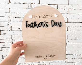May include: A wooden Father's Day sign with a light wood grain finish. The sign has an arched top with the words "our first Father's Day" in black lettering. Two small white hearts are on the top. The sign is personalized with "Nathan & Daddy 2024".