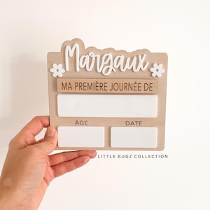 Könnte beinhalten: Ein Holzschild mit dem Namen "Margaux" in weißer Schrift, verziert mit Blumen. Das Schild enthält den Text "MA PREMIÈRE JOURNÉE DE" und Felder für Alter und Datum. Der Text "LITTLE BUGZ COLLECTION" befindet sich unten.