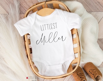 May include: White baby onesie with the text "Littlest Miller" in black script font. The onesie is in a wooden basket with a white blanket and brown shoes.