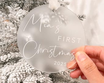 Könnte beinhalten: Ein klarer, runder Weihnachtsschmuck mit dem Text "Mia's FIRST Christmas 2025" in weißer Schrift. Der Schmuck wird vor einem gefrosteten Weihnachtsbaum gehalten. Die Worte "Little Bugz Collection" stehen oben.