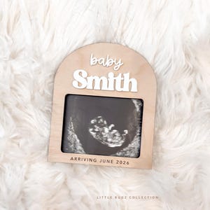 Op de afbeelding: Een lichtbruin houten frame met een echografie. Het frame heeft de witte woorden "baby Smith" bovenaan en "ARRIVING JUNE 2026" onderaan. Het frame heeft een boogvormige bovenkant en een rechthoekige opening.