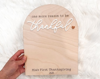 May include: Wooden sign with an arched top, featuring the words "one more reason to be thankful" in a script font. Below, it reads "Mia's First Thanksgiving 2025". A small heart is visible. The sign is made of light-colored wood.