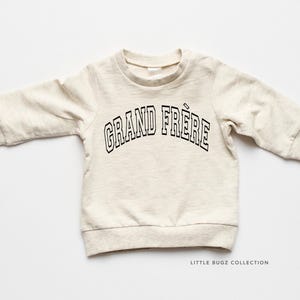 May include: A light beige baby sweatshirt with the words "GRAND FRÈRE" in black, arched lettering. The sweatshirt has long sleeves and a crew neck. The text is outlined in black. The words mean "big brother" in French.