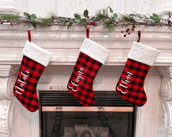 Puede incluir: Tres calcetines de Navidad de cuadros rojos y negros de búfalo con ribete de piel blanca colgados de una repisa de chimenea blanca. Los calcetines están personalizados con los nombres "Natalie", "Layna" y "Ethan" en letras blancas en cursiva.