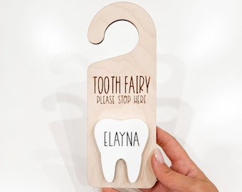 Puede incluir: Colgador de puerta de madera para la hada de los dientes con un recorte de diente blanco que dice "Elayna" y el texto "Tooth Fairy Please Stop Here". El colgador está hecho por Little Bugz Collection.