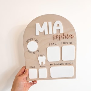 May include: A wooden milestone board with the name "MIA" in white letters and "sophia" in pink. It has sections for "Today I am Months Old," "I Can," "I Dislike," "I Have," "I Stand," "I Weigh," and "My Favorite Things."