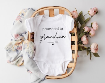 May include: White baby onesie with the text "promoted to grandma summer 2022" in black script. The onesie is inside a woven basket, with a floral scarf and pink flowers in the background.