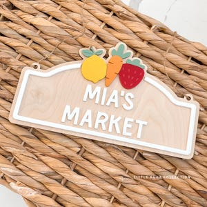 May include: Wooden sign with the text "MIA'S MARKET" in white, featuring a lemon, carrot, and strawberry. The sign has a white border and is hanging on a woven basket.