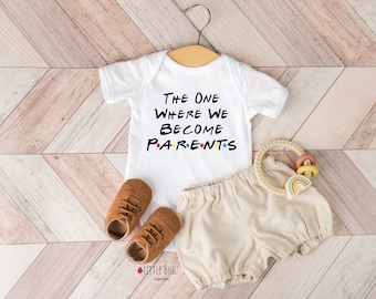 May include: White baby onesie with the text "The One Where We Become Parents" in a black font. The onesie is laid out on a wood floor with a pair of brown baby shoes and a beige baby romper.