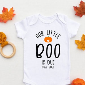 May include: White baby onesie with the text "OUR LITTLE BOO IS DUE MAY 2021" and a pumpkin graphic. The onesie is surrounded by fall-themed decorations, including orange and yellow leaves, a wooden teething ring, a small pumpkin, a pinecone, and a decorative gourd.