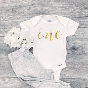 May include: White baby onesie with gold glitter lettering that says "one". The onesie is paired with gray pants.