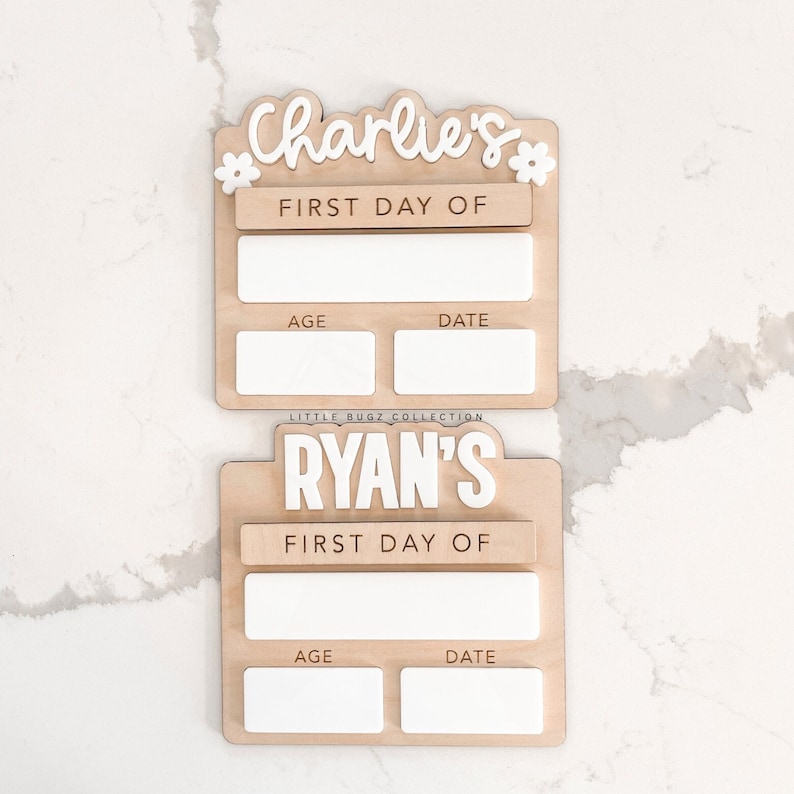 K&ouml;nnte beinhalten: Zwei Holzschildchen mit wei&szlig;en Buchstaben. Das obere Schildchen sagt "Charlie's" und das untere Schildchen sagt "Ryan's". Beide Schildchen haben den Text "First Day Of" und Platz f&uuml;r Alter und Datum.