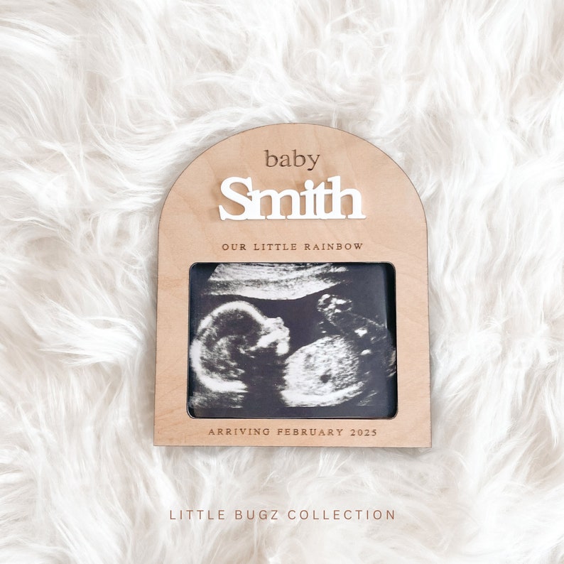May include: A wooden arch-shaped frame with a white ultrasound image inside. The frame has the text "baby Smith our little rainbow arriving February 2025" and "Little Bugz Collection" at the bottom.