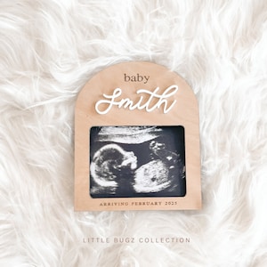 May include: A wooden frame with a white arch and the text "baby Smith" in white lettering. The frame has a black and white ultrasound image inside and the text "arriving February 2025". The frame is on a white fluffy surface and the text "LITTLE BUGZ COLLECTION" is below the frame.