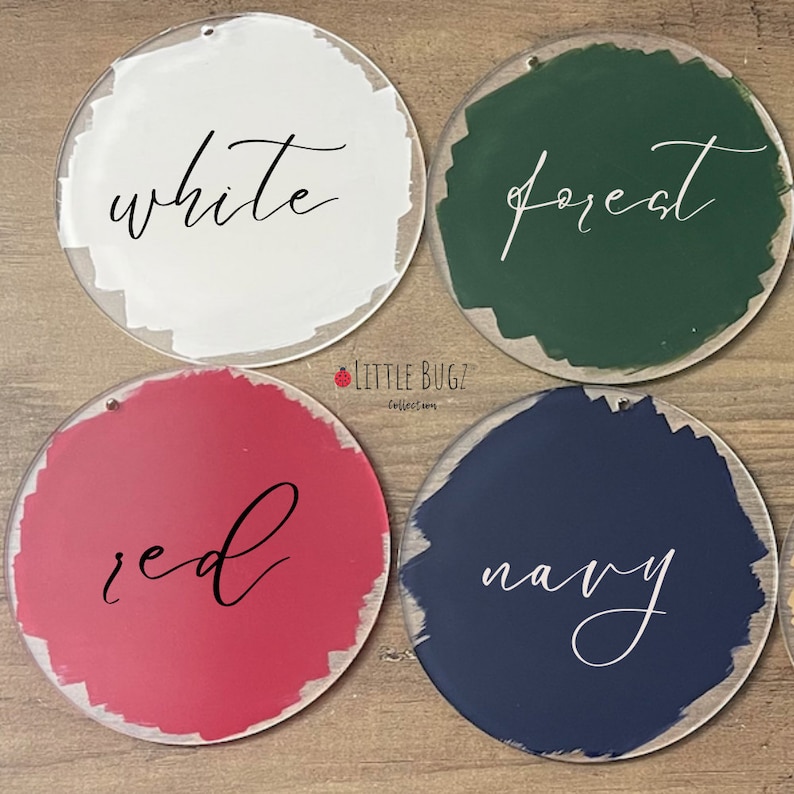 Puede incluir: Cuatro adornos redondos de acr&iacute;lico transparente con muestras de color pintadas en blanco, verde bosque, rojo y azul marino. Cada adorno tiene una palabra escrita a mano en cursiva que indica el color. Tambi&eacute;n es visible el logotipo de Little Bugz Collection.