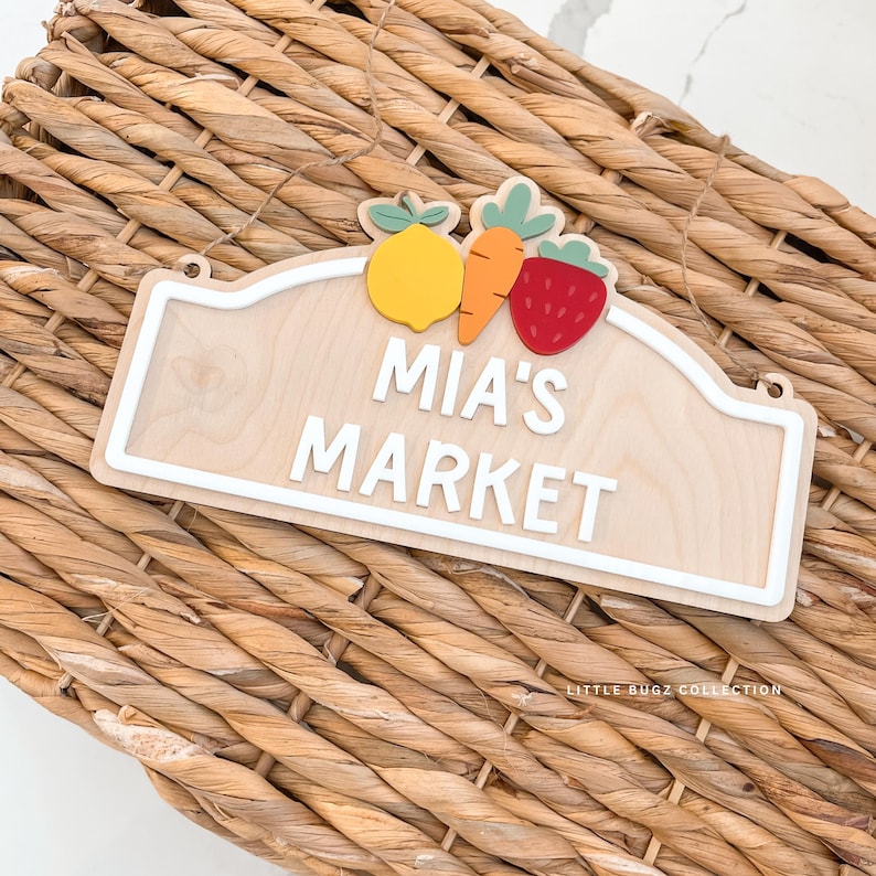 May include: Wooden sign with the text "MIA'S MARKET" in white, featuring a lemon, carrot, and strawberry. The sign has a white border and is hanging on a woven basket.