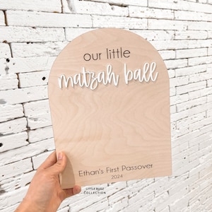 May include: A wooden sign with a rounded top, painted in a light wood colour. The sign says "our little matzah ball" in white cursive lettering. Below the text, it says "Ethan's First Passover 2024" in black lettering. The sign is held by a hand.