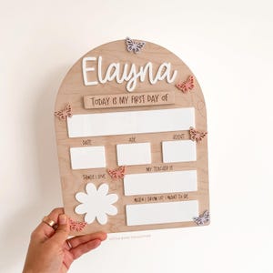 Op de afbeelding: Een boogvormig houten bord met de naam Elayna in wit schrift, met lege witte ruimtes voor het noteren van details over de eerste schooldag zoals datum, leeftijd, lengte, leraar en interesses, versierd met kleine pastelkleurige vlinders.