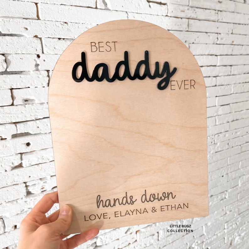 May include: Wooden sign with the text "Best daddy ever" in black lettering. The sign also has the text "hands down" and "Love, Elayna & Ethan" in black lettering. The sign is on a white brick background.
