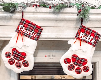 Puede incluir: Dos calcetines de Navidad de cuadros rojos y negros con huellas de patas blancas y los nombres "Charlie" y "Belle" bordados.