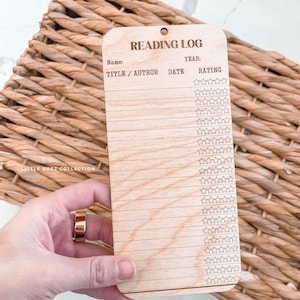 Peut inclure: Journal de lecture en bois avec le texte "Reading Log", "Name", "Year", "Title/Author", "Date" et "Rating". Le journal comporte des lignes pour l'écriture et des étoiles pour les notes. Le journal est brun clair et percé d'un trou en haut.