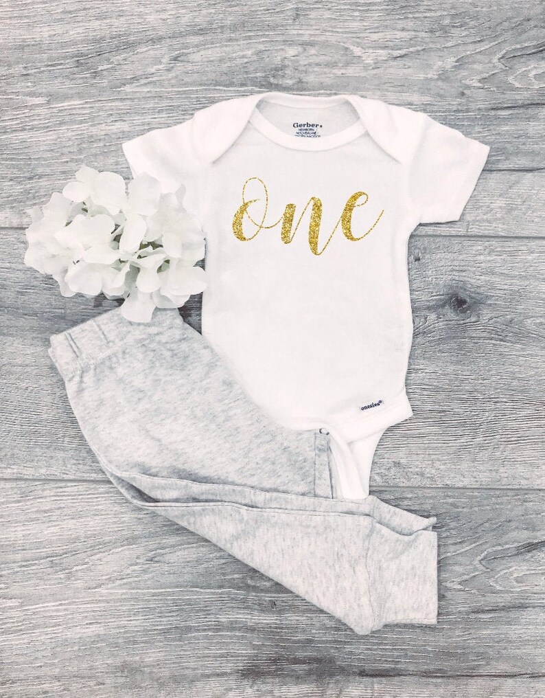 May include: White baby onesie with gold glitter lettering that says "one". The onesie is paired with gray pants.
