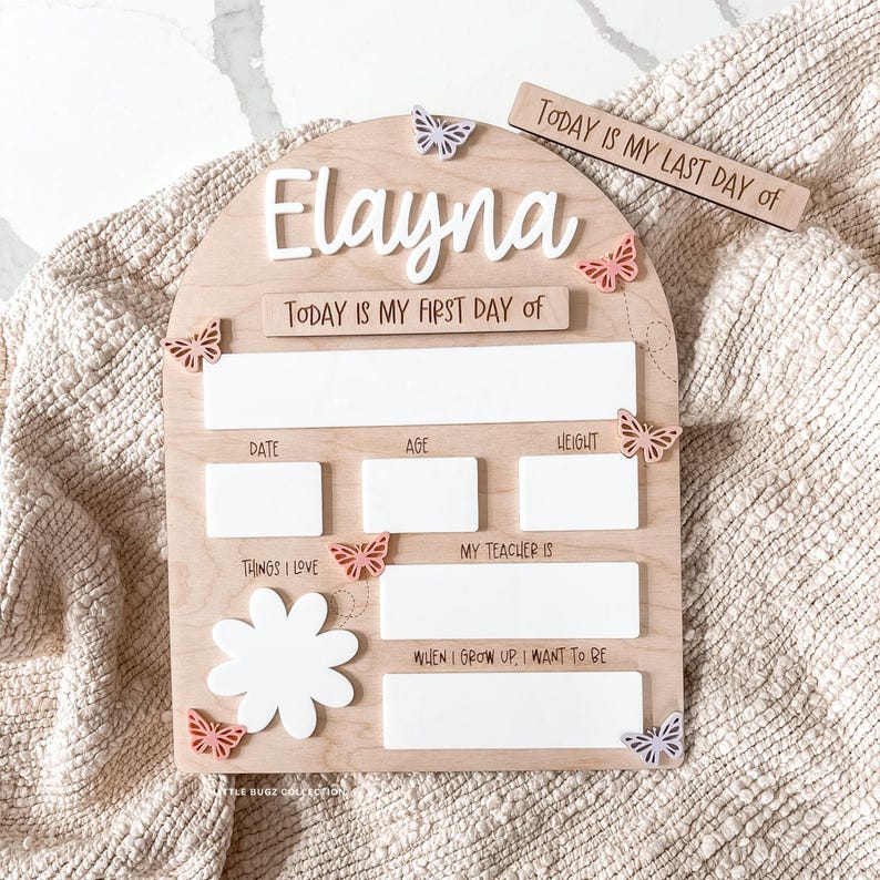 Op de afbeelding: Een boogvormig houten bord met de naam Elayna in wit schrift, voorzien van de teksten Today is my first day of, Date, Age, Height, Things I love, My teacher is en When I grow up, I want to be, versierd met kleine kleurrijke vlinders.