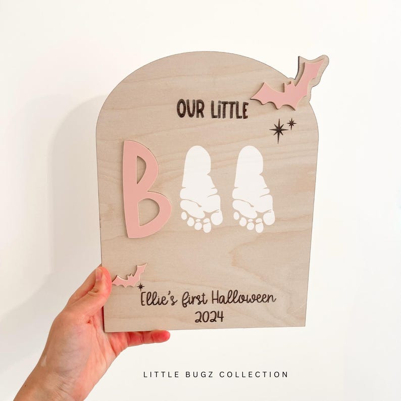 May include: A wooden sign with a pink "B" and two white footprints. The sign says "Our Little Boo" and "Ellie's first Halloween 2024". There are pink bats and stars on the sign.