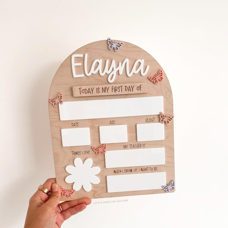 Op de afbeelding: Een boogvormig houten bord met de naam Elayna in wit handschrift. Het bevat de teksten Today is my first day of, Date, Age, Height, Things I love, My teacher is en When I grow up I want to be, met witte acryl invulvelden en vlinderaccenten.