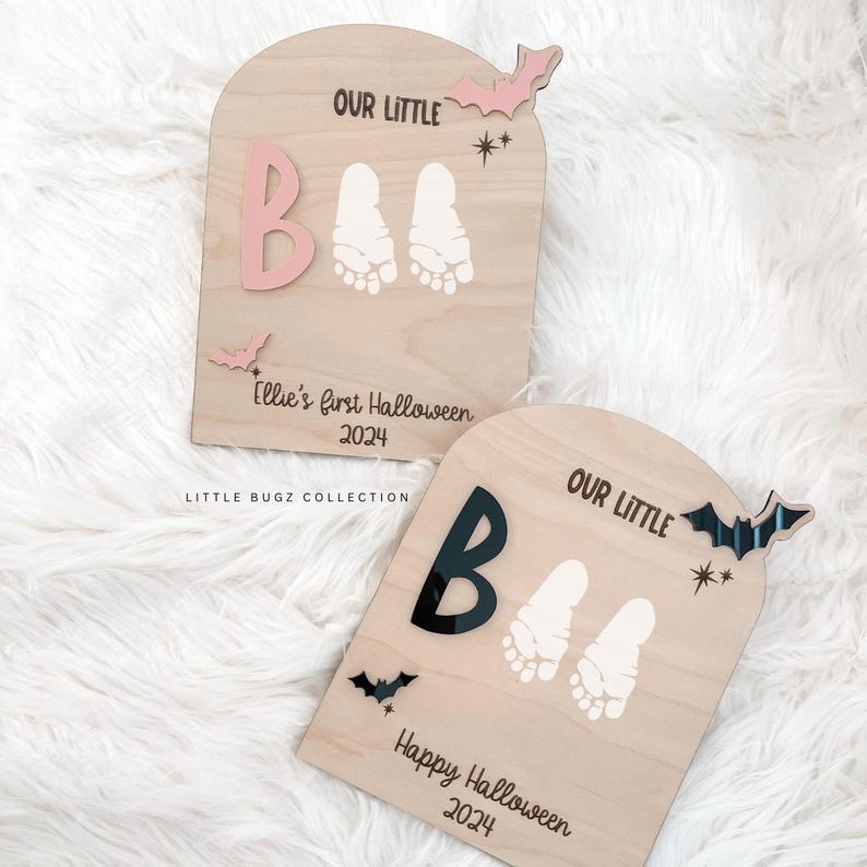 May include: Two wooden Halloween signs with the words "Our Little" and "Boo" in pink and black lettering. The signs feature a pair of baby footprints and a bat cutout. The first sign says "Ellie's first Halloween 2024" and the second sign says "Happy Halloween 2024".