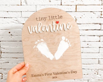 May include: A wooden sign with a white arch shape, the words "tiny little valentine" with a red heart, and two white handprints. The sign also has the text "Emma's First Valentine's Day 2024" and "Little Bugz Collection".