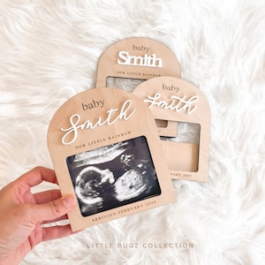 May include: Wooden photo frame with a white arch and the text "baby Smith our little rainbow" and a black and white ultrasound image. The frame has a space for a second photo and the text "arriving February 2025".