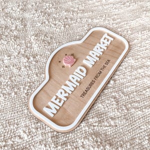 May include: A wooden sign with a white border featuring the raised white text MERMAID MARKET and TREASURES FROM THE SEA. A small pink seashell icon is positioned to the left of the text. The sign has a smooth light wood finish and rounded edges.