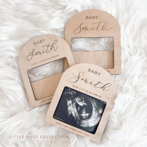 May include: Three wooden photo frames with arched tops. Each frame has a cutout for a photo and is engraved with the text "Baby Smith" and "Our Little Rainbow". The bottom frame has an ultrasound photo in the cutout and the text "Arriving February 2025".